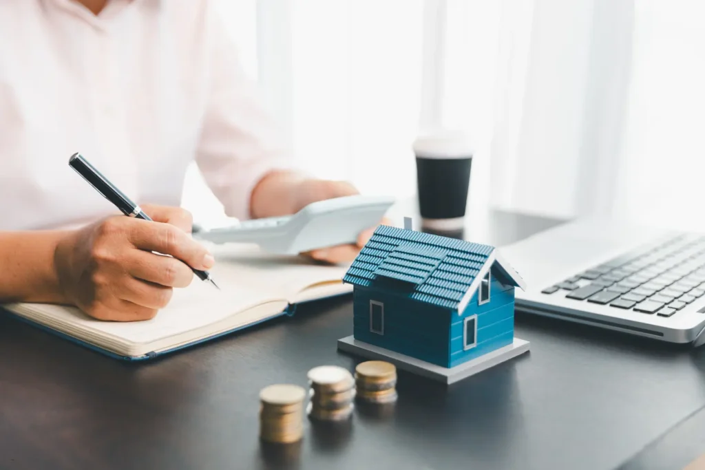 Property management tools shown with a person calculating finances beside a house model and stacked coins.