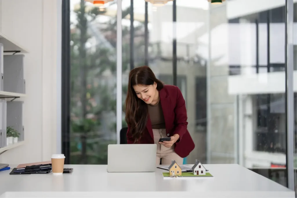 Tenant screening software represented by a real estate agent using a laptop and smartphone at her desk with house models.