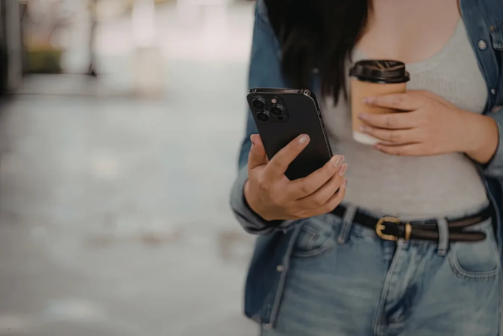 Property management app on smartphone held by woman drinking coffee outdoors in casual attire.