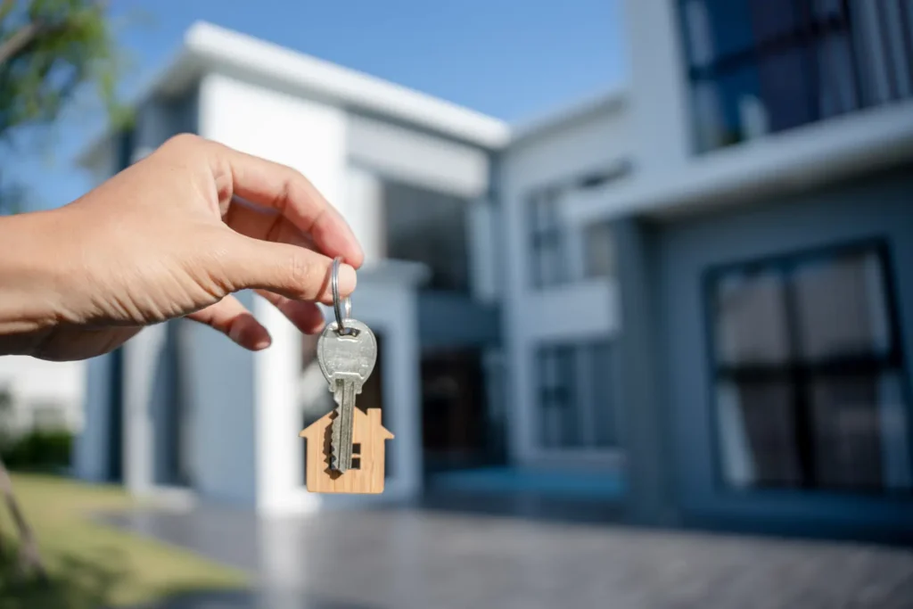Property management software pricing that helps landlords manage rentals illustrated by a hand holding a house key with a wooden home keychain in front of a modern two-story rental property