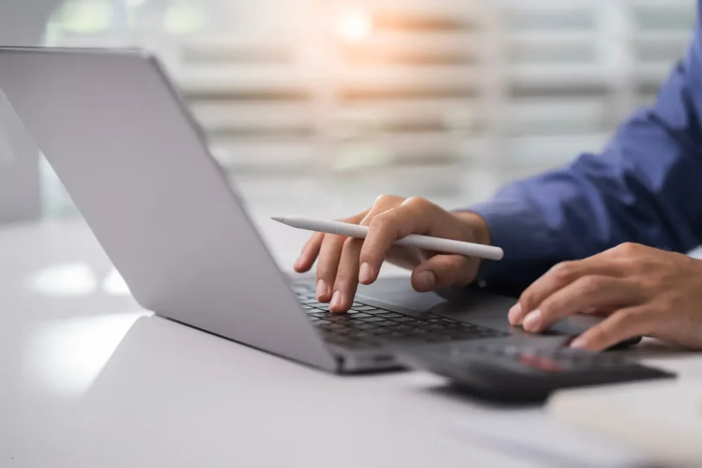 Tax deduction tracker app being used by a professional holding a stylus while typing on a laptop with a calculator at a bright office desk