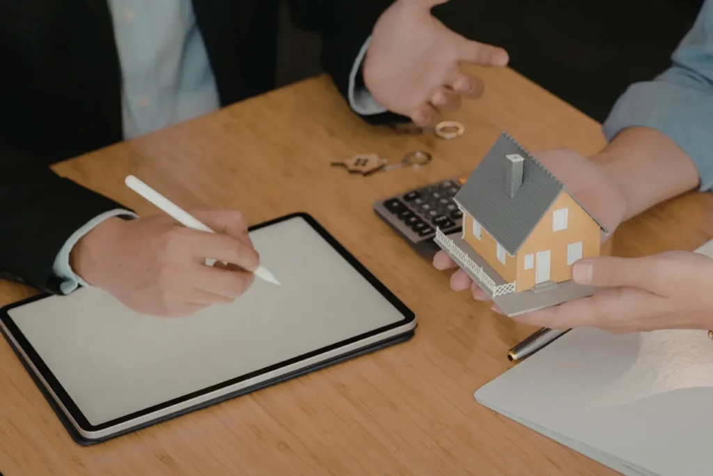 Mileage log for landlords supported by digital record-keeping as a real estate professional signs a tablet while a client holds a miniature house model across the desk